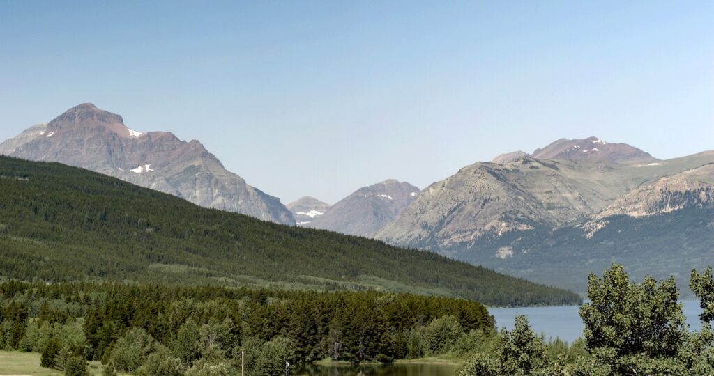 Rugged peaks with patches of snow above a forested ridge and a blue lake, green trees in the foreground