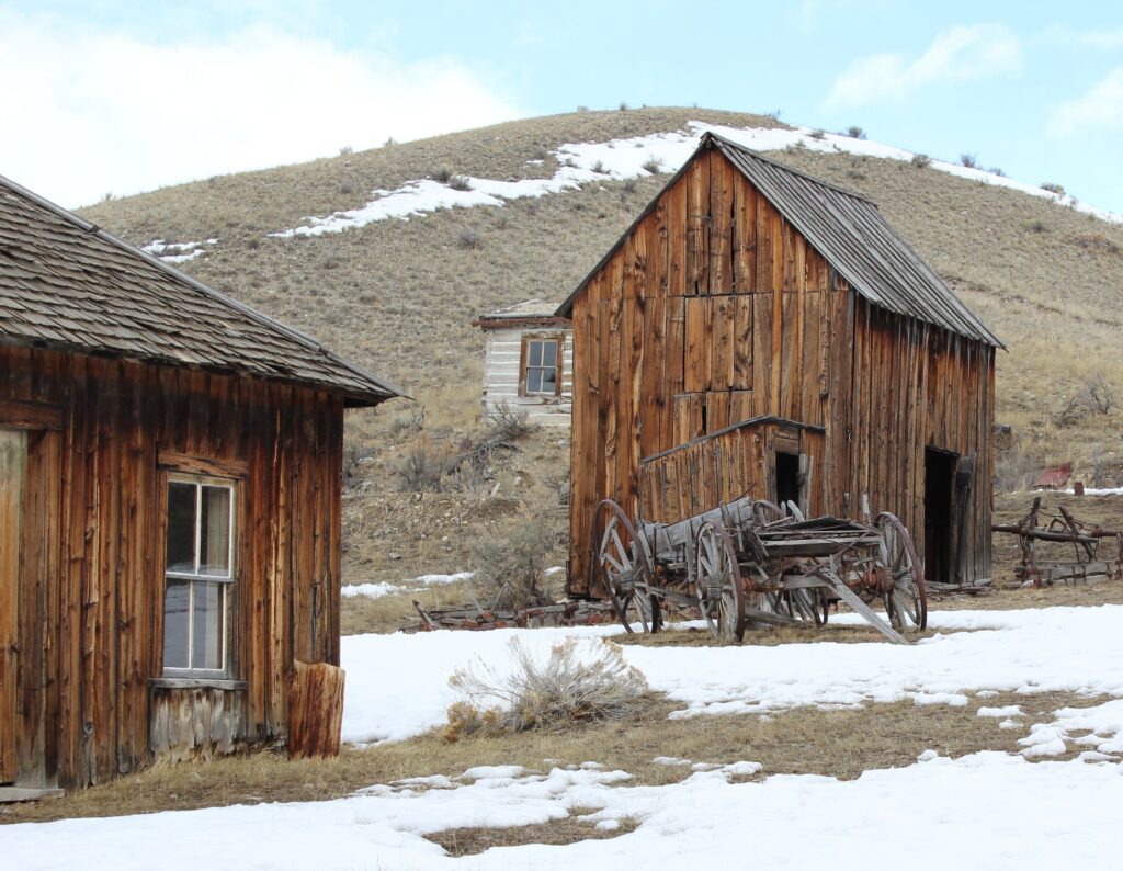 Weathered wooden buildings and an old wagon in the snow at Bannack State Park ghost town, Montana