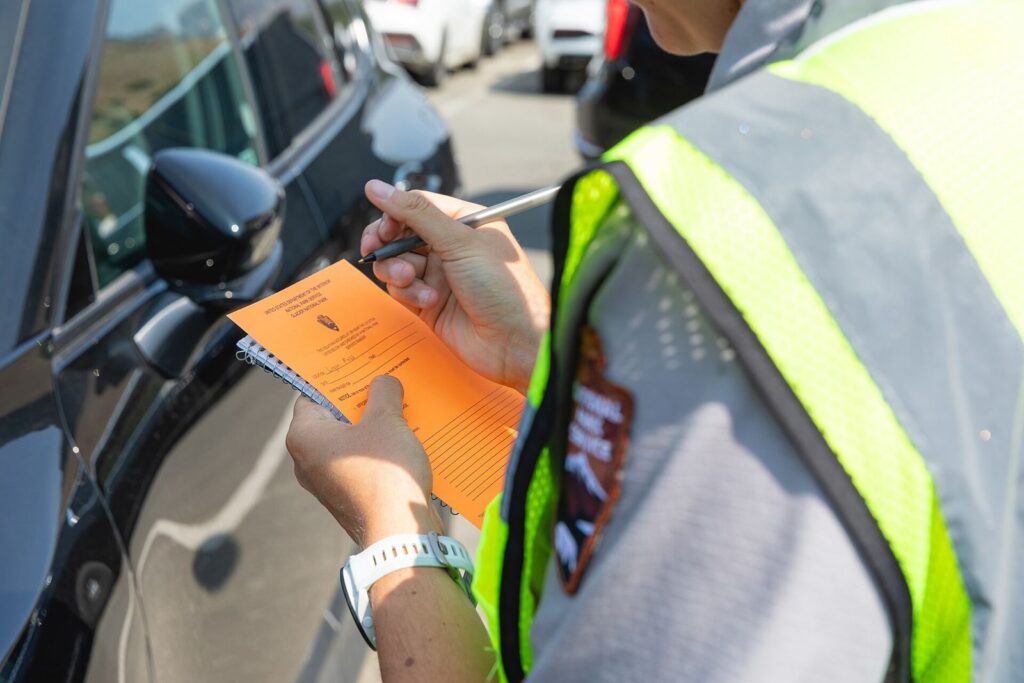 Parking attendant in a safety vest writing on a notepad beside a car in the Logan Pass parking lot