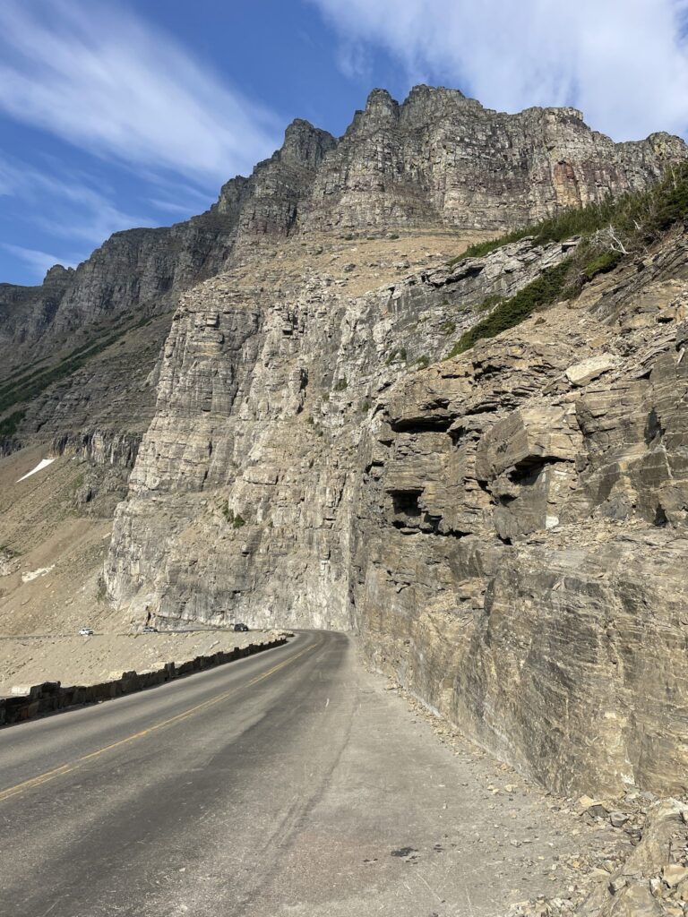Empty two-lane Going-to-the-Sun Road curving beneath a towering gray rock wall under a blue sky in Glacier National Park